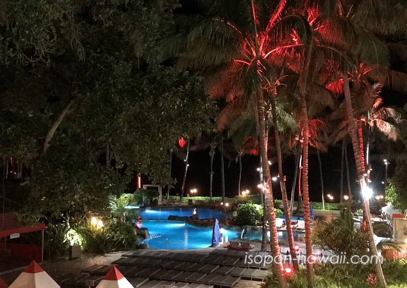 夜のヘルモア・プレイグラウンド。ヤシの木に赤い光が当たり、プールは青く輝いている。
